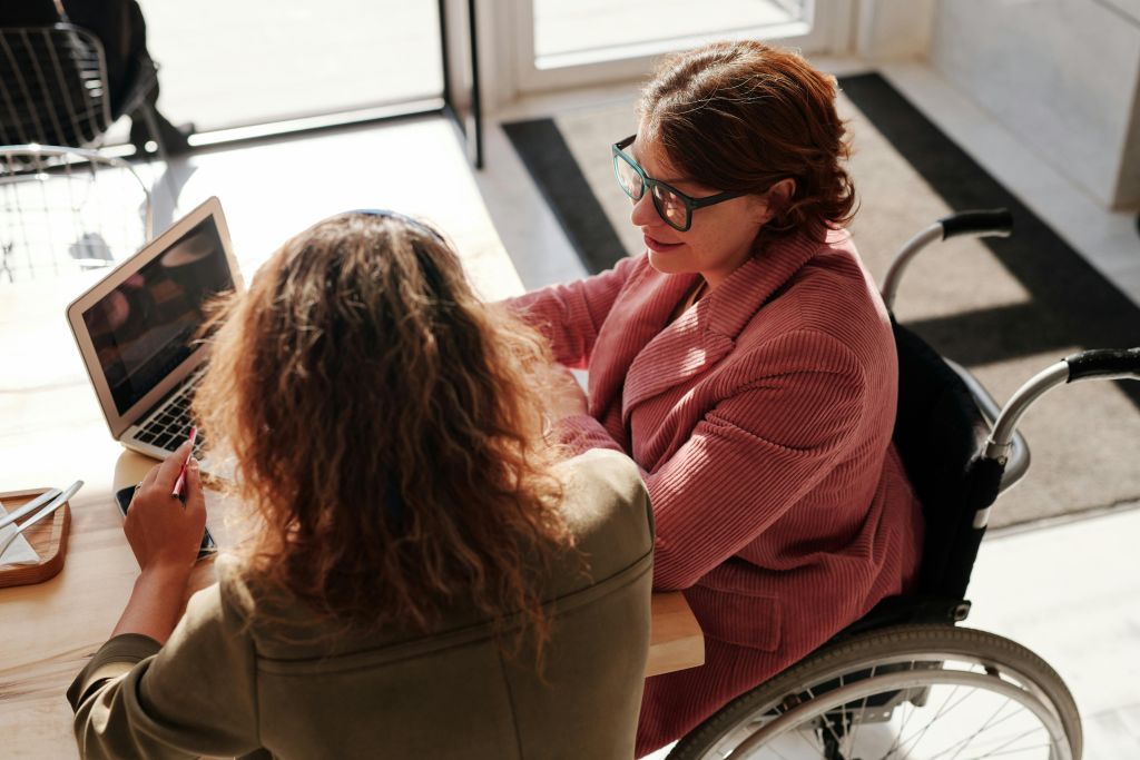 An overhead view of two women collaborating at a wooden table in a sunlit room. One woman, who has reddish-brown hair and glasses and uses a wheelchair, speaks with the other woman, who is seen from behind as she works on a laptop.