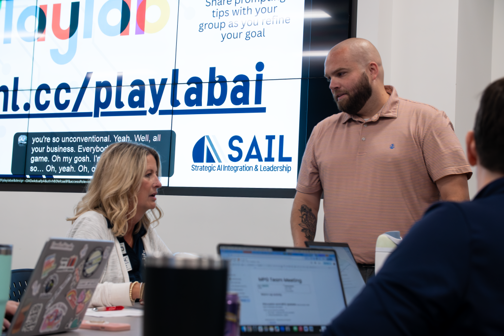 AS1 A man and a woman have a discussion in a professional meeting or workshop setting. The man, who is bald with a beard, stands and looks down at the seated blonde woman as she speaks. Laptops are open on the table in front of them. A large screen in the background displays information for an AI workshop, including the "SAIL: Strategic AI Integration & Leadership" logo.