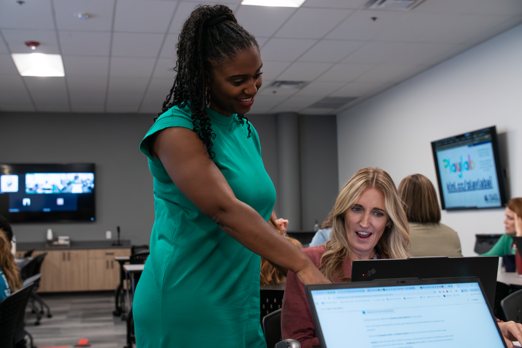 AS6 A woman in a green dress stands smiling and leans over to point at a laptop screen, assisting a woman who is seated at the desk. The seated woman looks at the screen with an engaged and surprised expression. They are in a modern office or training room setting.
