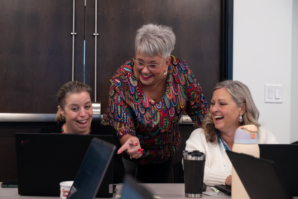 AS7 Three women share a laugh while gathered around laptops in an office or workshop setting. A standing woman with short gray hair and a brightly colored blouse leans over and points at a screen, smiling. Of the two seated women, one looks at the screen with a joyful, surprised expression while the other laughs.