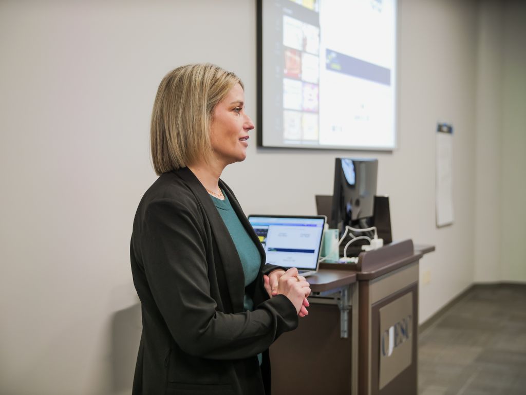 PGH67547-2 A woman with a blonde bob haircut, wearing a black blazer over a green shirt, stands at a podium giving a presentation. She is shown from the side, speaking, with her hands clasped in front of her. A laptop is on the podium, and a large projector screen is visible in the background.