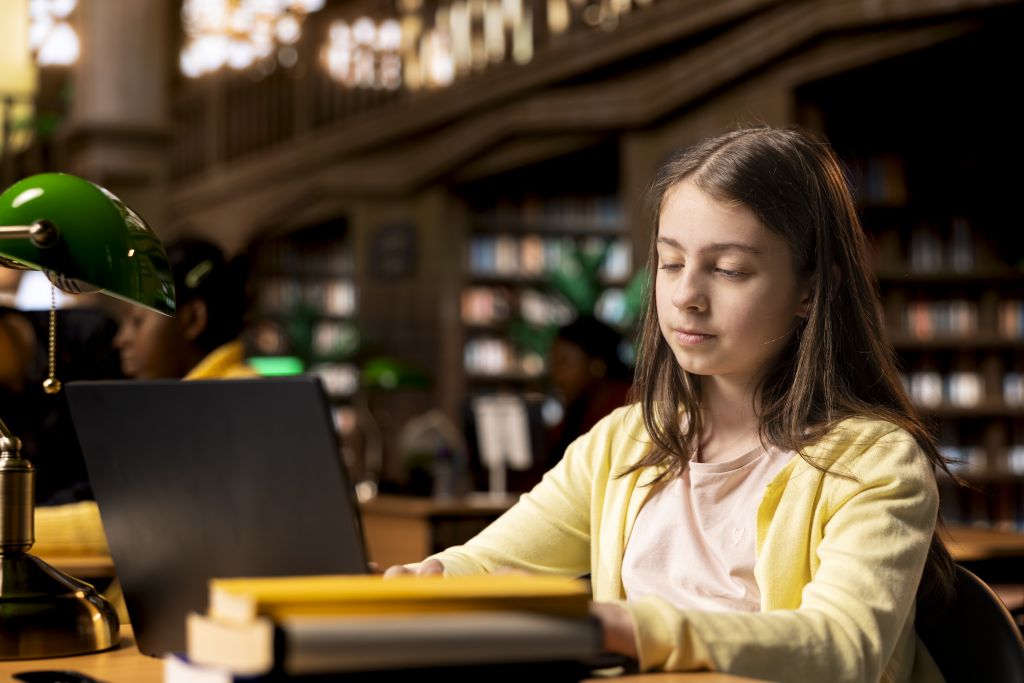 Young student competing her class notes on her laptop during a homework session, working on exercises in the school library. Teenager focusing on modern ways to finish assignments.